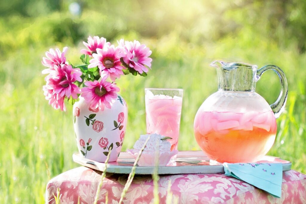 Vase mit pinken Blumen neben Karaffe und Glas mit rosa Getränk auf Tisch im Garten. Bild von Pixabay